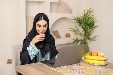 A quiet session at home, an Emirati woman performing remote work tasks, enjoying drinking Arabic coffee, integrating modern technology with her daily routine, a Saudi Arabian Gulf woman wearing an abaya and hijab sitting behind a table at home using a laptop, types of fresh fruits