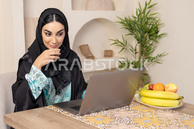 تعابير الاندماج والتركيز، سيدة إماراتية يقوم بأداء مهام العمل عن بعد، الاستمتاع بشرب القهوة العربية، دمج التكنولوجيا الحديثة مع الروتين اليومي، امرأة عربية خليجية سعودية ترتدي العباءة والحجاب تجلس خلف طاولة في المنزل تستخدم الحاسوب المحمول، أنواع الفاكهة الطازجة