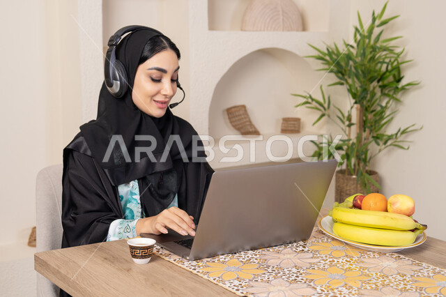 Performing work tasks remotely, enjoying drinking Arabic coffee, integrating modern technology with daily routine, healthy lifestyle, a Saudi Arabian Gulf woman wearing an abaya and hijab sitting at a table at home using a laptop, an Emirati woman wearing headphones, eating fresh fruit