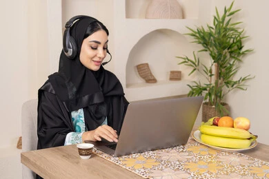 Performing work tasks remotely, enjoying drinking Arabic coffee, integrating modern technology with daily routine, healthy lifestyle, a Saudi Arabian Gulf woman wearing an abaya and hijab sitting at a table at home using a laptop, an Emirati woman wearing headphones, eating fresh fruit
