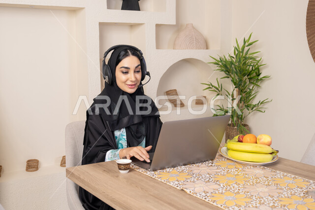 Performing work tasks remotely, enjoying drinking Arabic coffee, integrating modern technology with daily routine, healthy lifestyle, a Saudi Arabian Gulf woman wearing an abaya and hijab sitting at a table at home using a laptop, an Emirati woman wearing headphones, eating fresh fruit