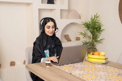 اداء مهام العمل عن بعد، الاستمتاع بشرب القهوة العربية، دمج التكنولوجيا الحديثة مع الروتين اليومي، نمط حياة صحي، امراه عربية خليجية سعودية ترتدي العباءة والحجاب تجلس على طاولة في المنزل تستخدم الحاسب المحمول، سيدة اماراتية تضع الهيدفون، تناول الفاكهة الطازجة