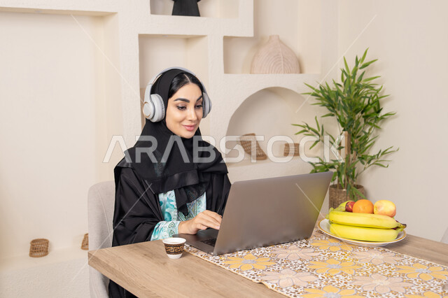 الاستمتاع بشرب القهوة العربية، دمج التكنولوجيا الحديثة مع الروتين اليومي، نمط حياة صحي، امراه عربية خليجية سعودية ترتدي العباءة والحجاب تجلس على طاولة في المنزل تستخدم الحاسب المحمول، سيدة اماراتية تضع الهيدفون، اداء مهام العمل عن بعد، تناول الفاكهة الطازجة