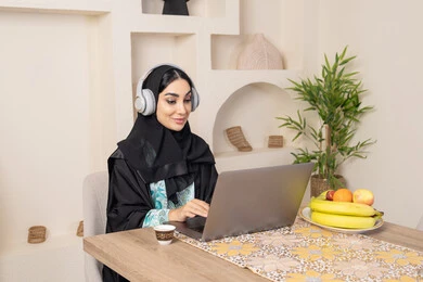 الاستمتاع بشرب القهوة العربية، دمج التكنولوجيا الحديثة مع الروتين اليومي، نمط حياة صحي، امراه عربية خليجية سعودية ترتدي العباءة والحجاب تجلس على طاولة في المنزل تستخدم الحاسب المحمول، سيدة اماراتية تضع الهيدفون، اداء مهام العمل عن بعد، تناول الفاكهة الطازجة