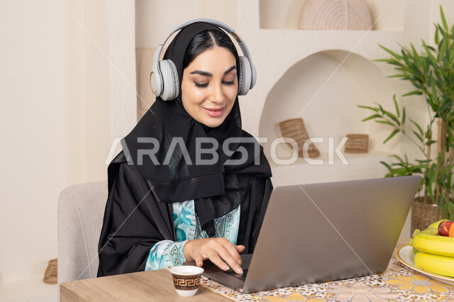 Healthy lifestyle, working remotely, enjoying Arabic coffee, integrating modern technology into daily routine, a Saudi Arabian Gulf woman wearing an abaya and hijab sitting at a table at home using a laptop, an Emirati woman wearing headphones