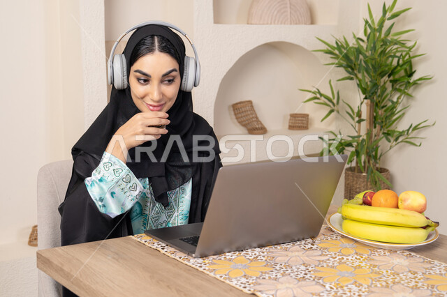 الاستمتاع بشرب القهوة العربية، دمج التكنولوجيا الحديثة مع الروتين اليومي، نمط حياة صحي، امراه عربية خليجية سعودية ترتدي العباءة والحجاب تجلس على طاولة في المنزل تستخدم الحاسب المحمول، سيدة اماراتية تضع الهيدفون، اداء مهام العمل عن بعد، تناول الفاكهة الطازجة