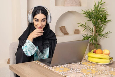الاستمتاع بشرب القهوة العربية، دمج التكنولوجيا الحديثة مع الروتين اليومي، نمط حياة صحي، امراه عربية خليجية سعودية ترتدي العباءة والحجاب تجلس على طاولة في المنزل تستخدم الحاسب المحمول، سيدة اماراتية تضع الهيدفون، اداء مهام العمل عن بعد، تناول الفاكهة الطازجة