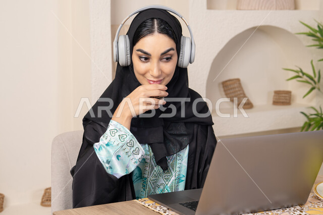 نمط حياة صحي، اداء مهام العمل عن بعد، الاستمتاع بشرب القهوة العربية، دمج التكنولوجيا الحديثة مع الروتين اليومي، امرأة عربية خليجية سعودية ترتدي العباءة والحجاب تجلس على طاولة في المنزل تستخدم الحاسب المحمول، سيدة اماراتية تضع الهيدفون