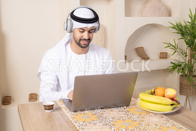 Fresh fruits and traditional Arabic coffee, integrating modern technology into daily routine, wearing earphones, working remotely, an Emirati Gulf Arab man wearing a kandura and ghutra sitting at a table at home working on his laptop, eating a healthy breakfast