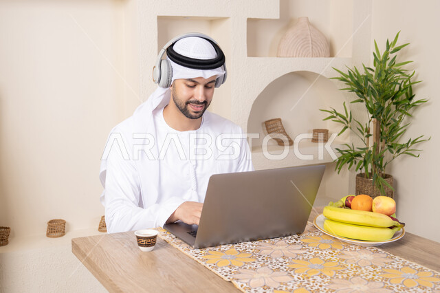 Wearing headphones, working remotely, an Emirati Gulf Arab man wearing a kandura and ghutra sitting at a table at home working on his laptop, eating a healthy breakfast, fresh fruits and traditional Arabic coffee, integrating modern technology with his daily routine.