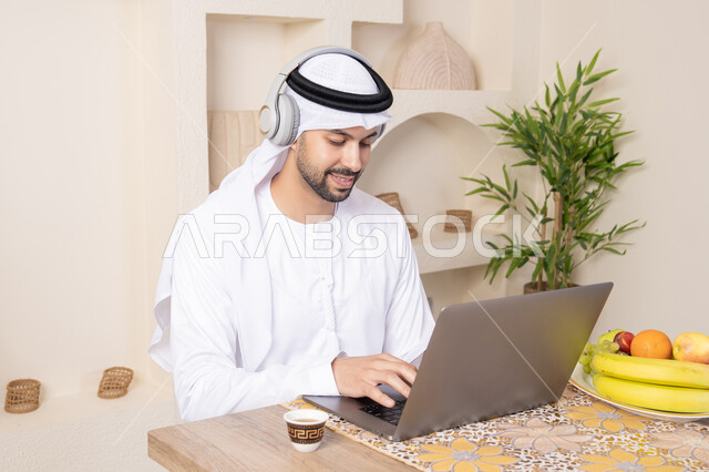 ارتداء سماعات الاذن، اداء مهام العمل عن بعد، رجل عربي خليجي إماراتي يرتدي الكندورة والغترة يجلس على طاولة في المنزل يعمل على الحاسب المحمول، تناول وجبة فطور صحية، أنواع الفاكهة الطازجة والقهوة العربية التقليدية، دمج التكنولوجيا الحديثة مع الروتين اليومي