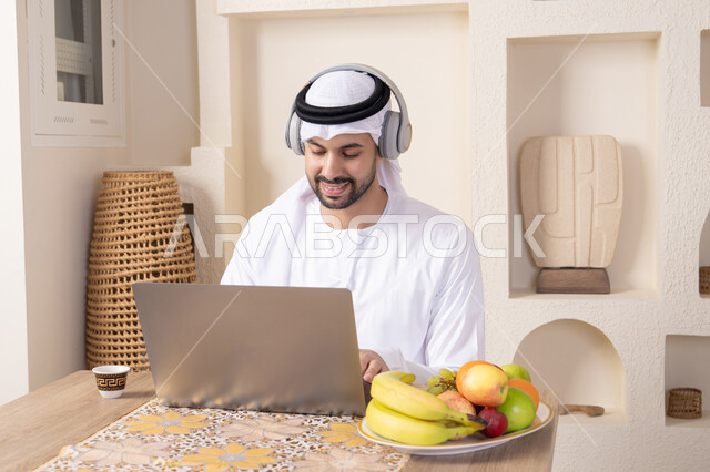 Fresh fruits and traditional Arabic coffee, integrating modern technology into daily routine, wearing earphones, working remotely, an Emirati Gulf Arab man wearing a kandura and ghutra sitting at a table at home working on his laptop, eating a healthy breakfast