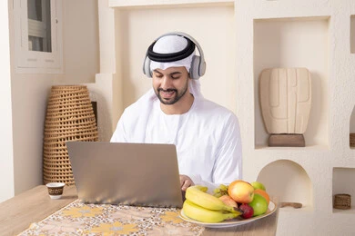 أنواع الفاكهة الطازجة والقهوة العربية التقليدية، دمج التكنولوجيا الحديثة مع الروتين اليومي، ارتداء سماعات الاذن، اداء مهام العمل عن بعد، رجل عربي خليجي إماراتي يرتدي الكندورة والغترة يجلس على طاولة في المنزل يعمل على الحاسب المحمول، تناول وجبة فطور صحية