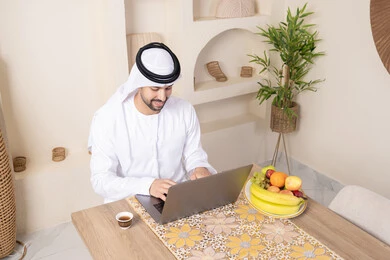 اداء مهام العمل عن بعد، رجل عربي خليجي إماراتي يرتدي الكندورة والغترة يجلس على طاولة في المنزل يعمل على الحاسب المحمول، تناول وجبة فطور صحية، أنواع الفاكهة الطازجة والقهوة العربية التقليدية، دمج التكنولوجيا الحديثة مع الروتين اليومي، نمط حياة صحي