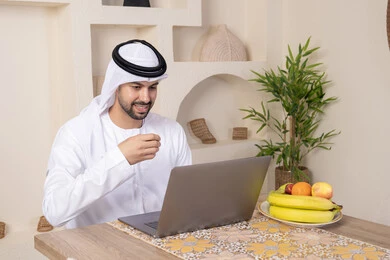 اداء مهام العمل عن بعد، رجل عربي خليجي إماراتي يرتدي الكندورة والغترة يجلس على طاولة في المنزل يعمل على الحاسب المحمول، تناول وجبة فطور صحية، أنواع الفاكهة الطازجة والقهوة العربية التقليدية، دمج التكنولوجيا الحديثة مع الروتين اليومي، نمط حياة صحي