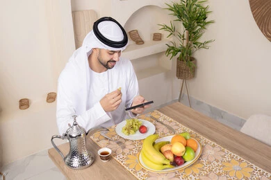 تناول وجبة فطور صحية، أنواع الفاكهة الطازجة والقهوة العربية التقليدية، دمج التكنولوجيا الحديثة مع الروتين اليومي، نمط حياة صحي، أجواء صباحية هادئة، رجل عربي خليجي إماراتي يرتدي الكندورة والغترة يجلس على طاولة في المنزل يستخدم الهاتف المحمول