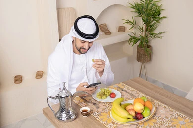 Calm morning atmosphere, an Emirati Gulf Arab man wearing a kandura and ghutra sitting at a table at home using a mobile phone, eating a healthy breakfast, fresh fruits and traditional Arabic coffee, integrating modern technology with daily routine, a healthy lifestyle