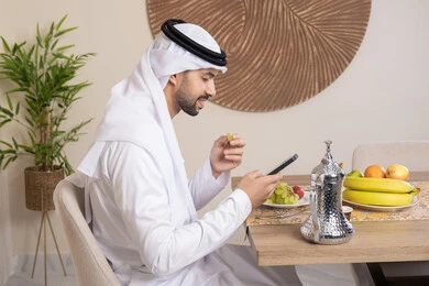 أنواع الفاكهة الطازجة والقهوة العربية التقليدية، دمج التكنولوجيا الحديثة مع الروتين اليومي، نمط حياة صحي، أجواء صباحية هادئة، رجل عربي خليجي إماراتي يرتدي الكندورة والغترة يجلس على طاولة في المنزل يستخدم الهاتف المحمول، تناول وجبة فطور صحية