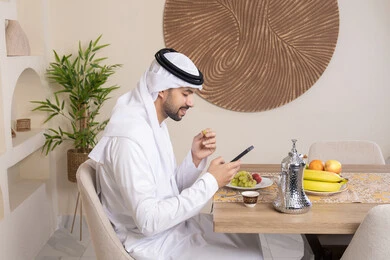 Eating a healthy breakfast, fresh fruits and traditional Arabic coffee, integrating modern technology with daily routine, healthy lifestyle, calm morning atmosphere, Emirati Gulf Arab man wearing kandura and ghutra sitting at a table at home using mobile phone