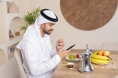 نمط حياة صحي، أجواء صباحية هادئة، رجل عربي خليجي إماراتي يرتدي الكندورة والغترة يجلس على طاولة في المنزل يستخدم الهاتف المحمول، تناول وجبة فطور صحية، أنواع الفاكهة الطازجة والقهوة العربية التقليدية، دمج التكنولوجيا الحديثة مع الروتين اليومي