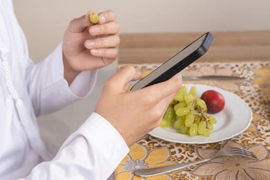 أنواع الفاكهة الطازجة والقهوة العربية التقليدية، دمج التكنولوجيا الحديثة مع الروتين اليومي، نمط حياة صحي، أجواء صباحية هادئة، رجل عربي خليجي إماراتي يرتدي الكندورة والغترة يجلس على طاولة في المنزل يستخدم الهاتف المحمول، تناول وجبة فطور صحية