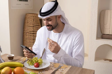نمط حياة صحي، أجواء صباحية هادئة، رجل عربي خليجي إماراتي يرتدي الكندورة والغترة يجلس على طاولة في المنزل يستخدم الهاتف المحمول، تناول وجبة فطور صحية، أنواع الفاكهة الطازجة والقهوة العربية التقليدية، دمج التكنولوجيا الحديثة مع الروتين اليومي