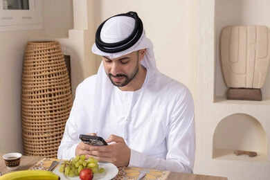 أجواء صباحية هادئة، رجل عربي خليجي إماراتي يرتدي الكندورة والغترة يجلس على طاولة في المنزل يستخدم الهاتف المحمول، تناول وجبة فطور صحية، أنواع الفاكهة الطازجة والقهوة العربية التقليدية، دمج التكنولوجيا الحديثة مع الروتين اليومي، نمط حياة صحي