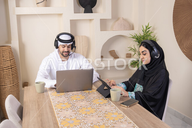 تناول المشروبات الساخنة، العمل عن بعد، الاستماع عبر الهيدفون، إنجاز المهام من المنزل، دمج التقنية والتكنولوجيا في الروتين اليومي، زوجان عربيان خليجيان اماراتيان يجلسان في البيت يعملان على الحاسب المحمول وجهاز التابلت، تعابير التركيز والسرور