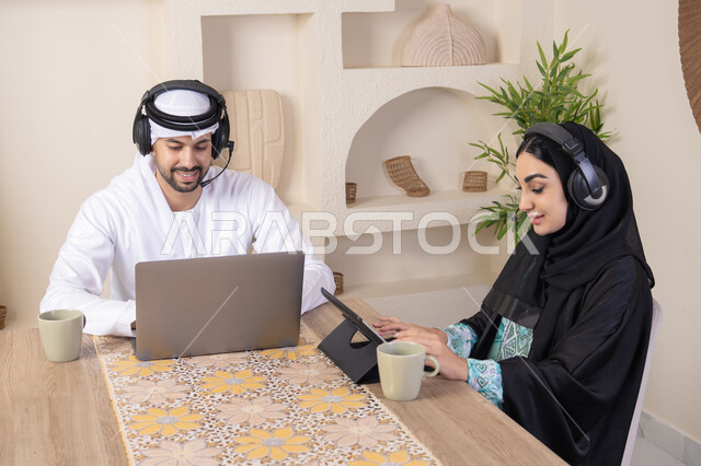 تناول المشروبات الساخنة، العمل عن بعد، الاستماع عبر الهيدفون، إنجاز المهام من المنزل، دمج التقنية والتكنولوجيا في الروتين اليومي، زوجان عربيان خليجيان اماراتيان يجلسان في البيت يعملان على الحاسب المحمول وجهاز التابلت، تعابير التركيز والسرور