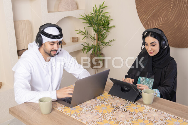 تناول المشروبات الساخنة، العمل عن بعد، الاستماع عبر الهيدفون، إنجاز المهام من المنزل، دمج التقنية والتكنولوجيا في الروتين اليومي، زوجان عربيان خليجيان اماراتيان يجلسان في البيت يعملان على الحاسب المحمول وجهاز التابلت، تعابير التركيز والسرور