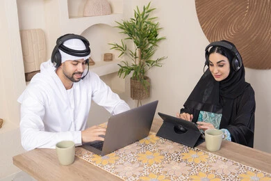 تناول المشروبات الساخنة، العمل عن بعد، الاستماع عبر الهيدفون، إنجاز المهام من المنزل، دمج التقنية والتكنولوجيا في الروتين اليومي، زوجان عربيان خليجيان اماراتيان يجلسان في البيت يعملان على الحاسب المحمول وجهاز التابلت، تعابير التركيز والسرور