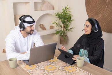 Listening on headphones, getting things done from home, incorporating technology into daily routine, an Emirati Gulf Arab couple sitting at home working on a laptop and tablet, expressions of concentration and pleasure, drinking hot drinks, working remotely