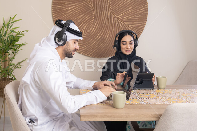 تعابير التركيز والسرور، تناول المشروبات الساخنة، العمل عن بعد، الاستماع عبر الهيدفون، إنجاز المهام من المنزل، دمج التقنية والتكنولوجيا في الروتين اليومي، زوجان عربيان خليجيان اماراتيان يجلسان في البيت يعملان على الحاسب المحمول وجهاز التابلت