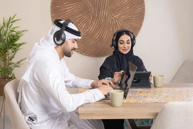 تعابير التركيز والسرور، تناول المشروبات الساخنة، العمل عن بعد، الاستماع عبر الهيدفون، إنجاز المهام من المنزل، دمج التقنية والتكنولوجيا في الروتين اليومي، زوجان عربيان خليجيان اماراتيان يجلسان في البيت يعملان على الحاسب المحمول وجهاز التابلت