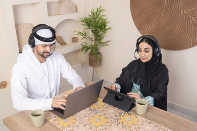 Listening on headphones, getting things done from home, incorporating technology into daily routine, an Emirati Gulf Arab couple sitting at home working on a laptop and tablet, expressions of concentration and pleasure, drinking hot drinks, working remotely