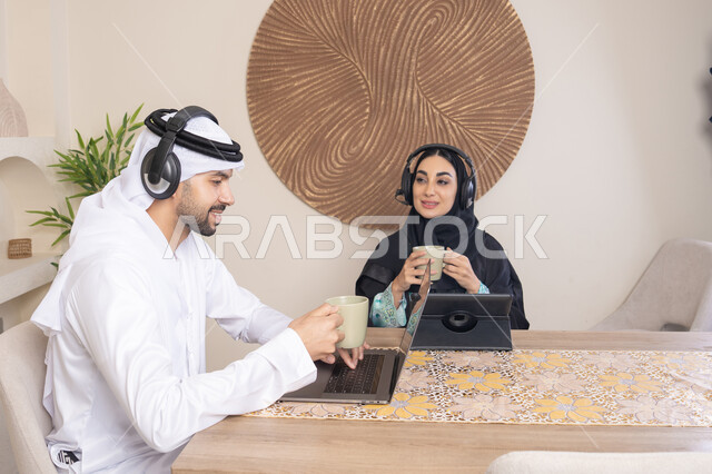 Expressions of focus and pleasure, drinking hot drinks, working remotely, listening to headphones, completing tasks from home, incorporating technology into daily routine, an Arab Gulf Emirati couple sitting at home working on a laptop and tablet