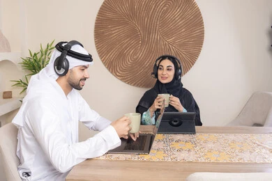Expressions of focus and pleasure, drinking hot drinks, working remotely, listening to headphones, completing tasks from home, incorporating technology into daily routine, an Arab Gulf Emirati couple sitting at home working on a laptop and tablet