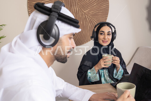 تعابير التركيز والسرور، تناول المشروبات الساخنة، العمل عن بعد، الاستماع عبر الهيدفون، إنجاز المهام من المنزل، دمج التقنية والتكنولوجيا في الروتين اليومي، زوجان عربيان خليجيان اماراتيان يجلسان في البيت يعملان على الحاسب المحمول وجهاز التابلت