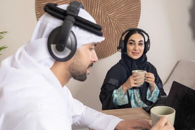 Expressions of focus and pleasure, drinking hot drinks, working remotely, listening to headphones, completing tasks from home, incorporating technology into daily routine, an Arab Gulf Emirati couple sitting at home working on a laptop and tablet