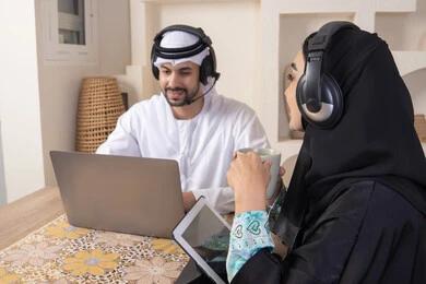 ايماءات التركيز والسرور، تناول المشروبات الساخنة، العمل عن بعد، الاستماع عبر الهيدفون، إنجاز المهام من المنزل، دمج التقنية والتكنولوجيا في الروتين اليومي، زوجان عربيان خليجيان اماراتيان يجلسان في البيت يعملان على الحاسب المحمول وجهاز التابلت