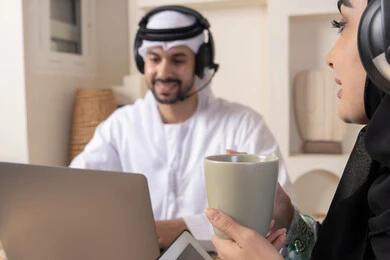 Listening on headphones, getting things done from home, incorporating technology into daily routine, an Emirati Gulf Arab couple sitting at home working on a laptop and tablet, expressions of concentration and pleasure, drinking hot drinks, working remotely