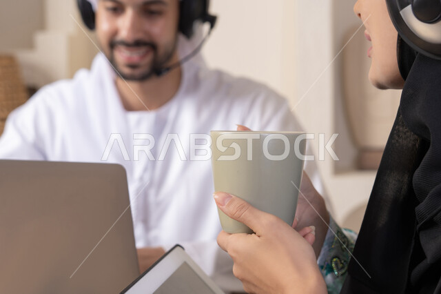 تعابير التركيز والسرور، تناول المشروبات الساخنة، العمل عن بعد، الاستماع عبر الهيدفون، إنجاز المهام من المنزل، دمج التقنية والتكنولوجيا في الروتين اليومي، زوجان عربيان خليجيان اماراتيان يجلسان في البيت يعملان على الحاسب المحمول وجهاز التابلت