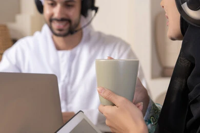 Expressions of focus and pleasure, drinking hot drinks, working remotely, listening to headphones, completing tasks from home, incorporating technology into daily routine, an Arab Gulf Emirati couple sitting at home working on a laptop and tablet
