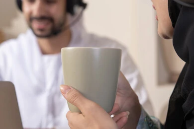 Listening on headphones, getting things done from home, incorporating technology into daily routine, an Emirati Gulf Arab couple sitting at home working on a laptop and tablet, expressions of concentration and pleasure, drinking hot drinks, working remotely
