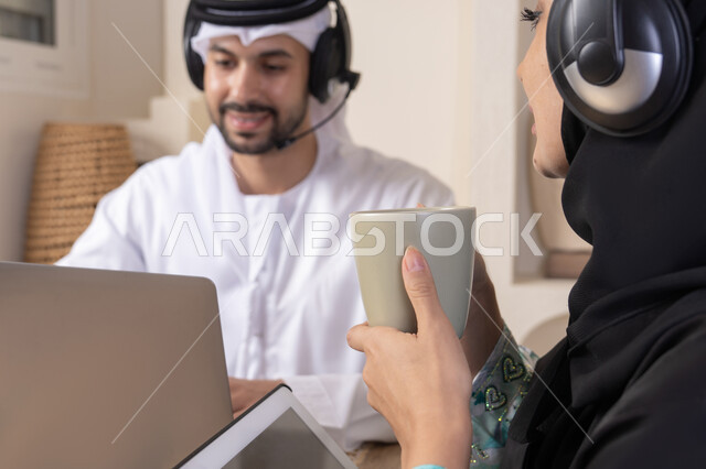 ايماءات التركيز والسرور، تناول المشروبات الساخنة، العمل عن بعد، الاستماع عبر الهيدفون، إنجاز المهام من المنزل، دمج التقنية والتكنولوجيا في الروتين اليومي، زوجان عربيان خليجيان اماراتيان يجلسان في البيت يعملان على الحاسب المحمول وجهاز التابلت