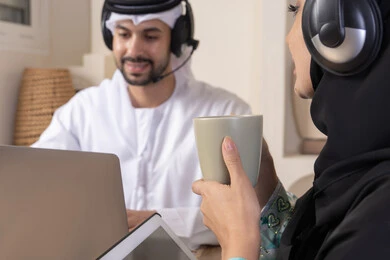 ايماءات التركيز والسرور، تناول المشروبات الساخنة، العمل عن بعد، الاستماع عبر الهيدفون، إنجاز المهام من المنزل، دمج التقنية والتكنولوجيا في الروتين اليومي، زوجان عربيان خليجيان اماراتيان يجلسان في البيت يعملان على الحاسب المحمول وجهاز التابلت