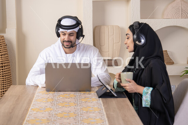 ايماءات التركيز والسرور، تناول المشروبات الساخنة، العمل عن بعد، الاستماع عبر الهيدفون، إنجاز المهام من المنزل، دمج التقنية والتكنولوجيا في الروتين اليومي، زوجان عربيان خليجيان اماراتيان يجلسان في البيت يعملان على الحاسب المحمول وجهاز التابلت