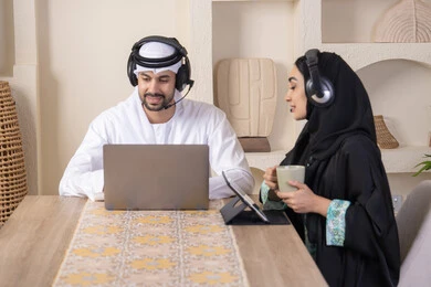 Gestures of focus and pleasure, drinking hot drinks, working remotely, listening on headphones, completing tasks from home, integrating technology into the daily routine, an Arab Gulf Emirati couple sitting at home working on a laptop and tablet