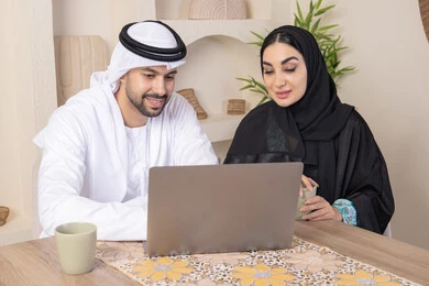Getting things done from home, incorporating technology into daily routine, an Emirati Gulf Arab couple sitting at home working on a laptop, gestures and expressions of focus and pleasure, drinking hot drinks, family bonding and communication, pointing at the laptop, working remotely