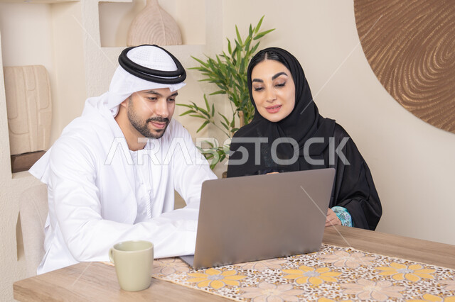 Getting things done from home, incorporating technology into daily routine, an Emirati Gulf Arab couple sitting at home working on a laptop, gestures and expressions of focus and pleasure, drinking hot drinks, family bonding and communication, pointing at the laptop, working remotely
